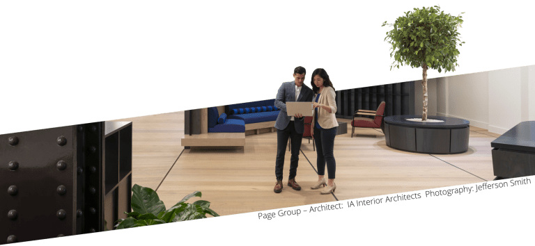 At Page Group Leeds, two professionals stand in their new office discussing at a laptop. The setting features stylish, contemporary furniture with vibrant blue and red accents and a large potted tree, contributing to the sophisticated yet welcoming atmosphere of the space.